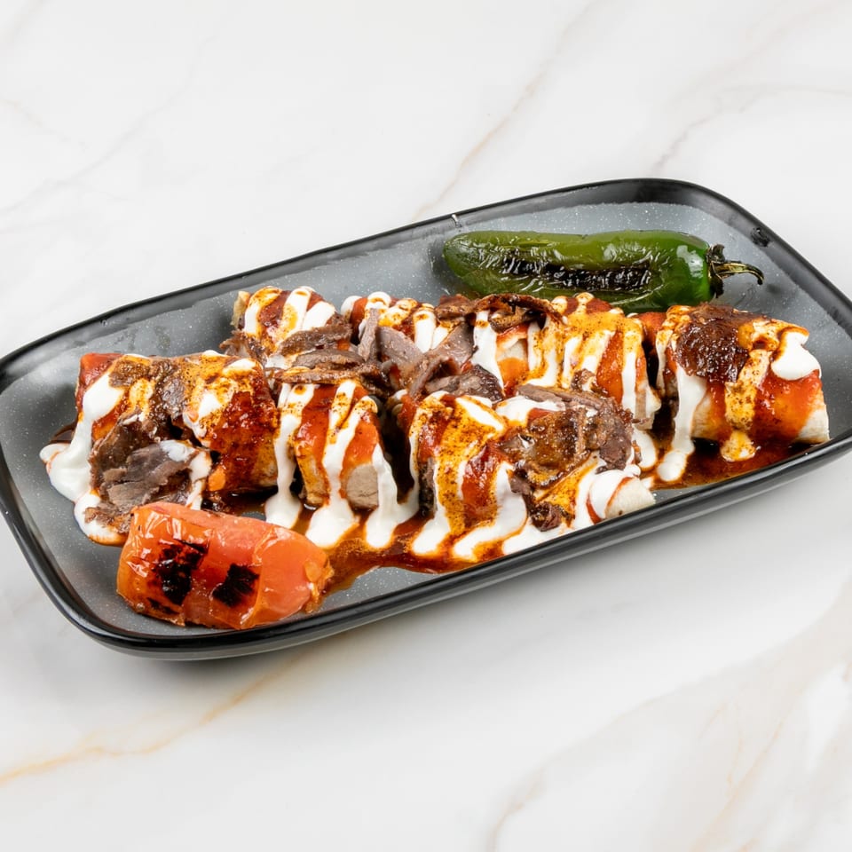 Marash Mediterranean Iskender kebab with yogurt, tomato sauce, grilled pepper, and tomato on a black plate.