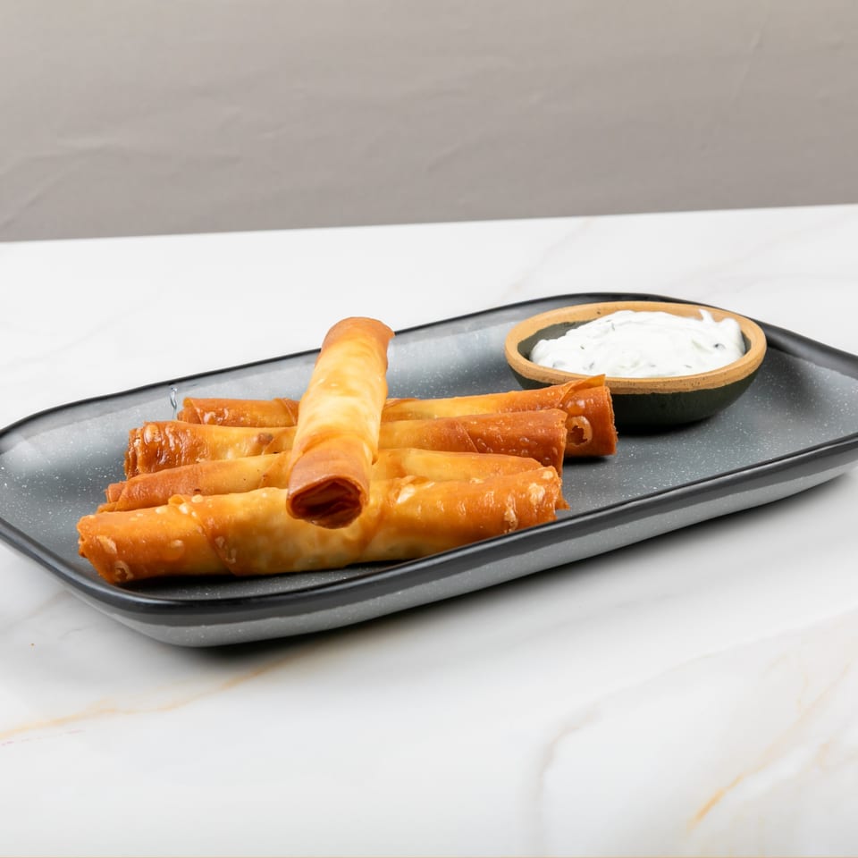 Golden-brown fried cheese rolls, a Mediterranean appetizer, served with a side of white dipping sauce.