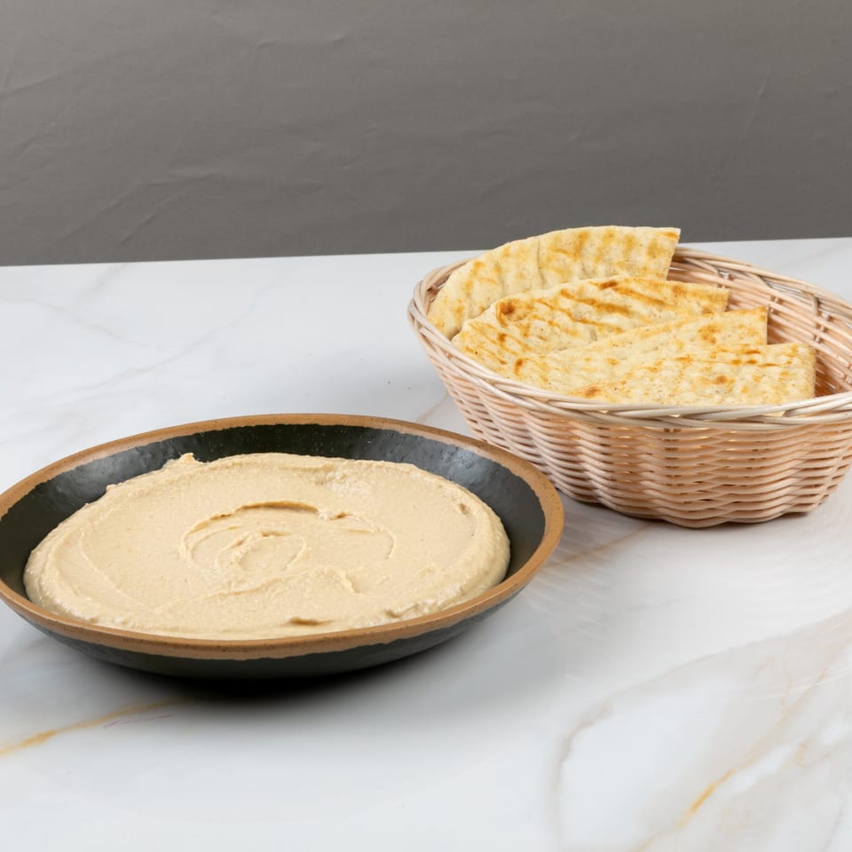 Creamy hummus in a black bowl with a basket of pita bread on a white marble surface.
